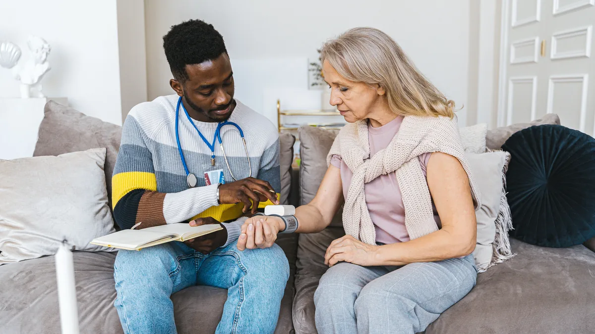 A health worker checks an older woman's blood pressure with an arm cuff while she sits on a sofa