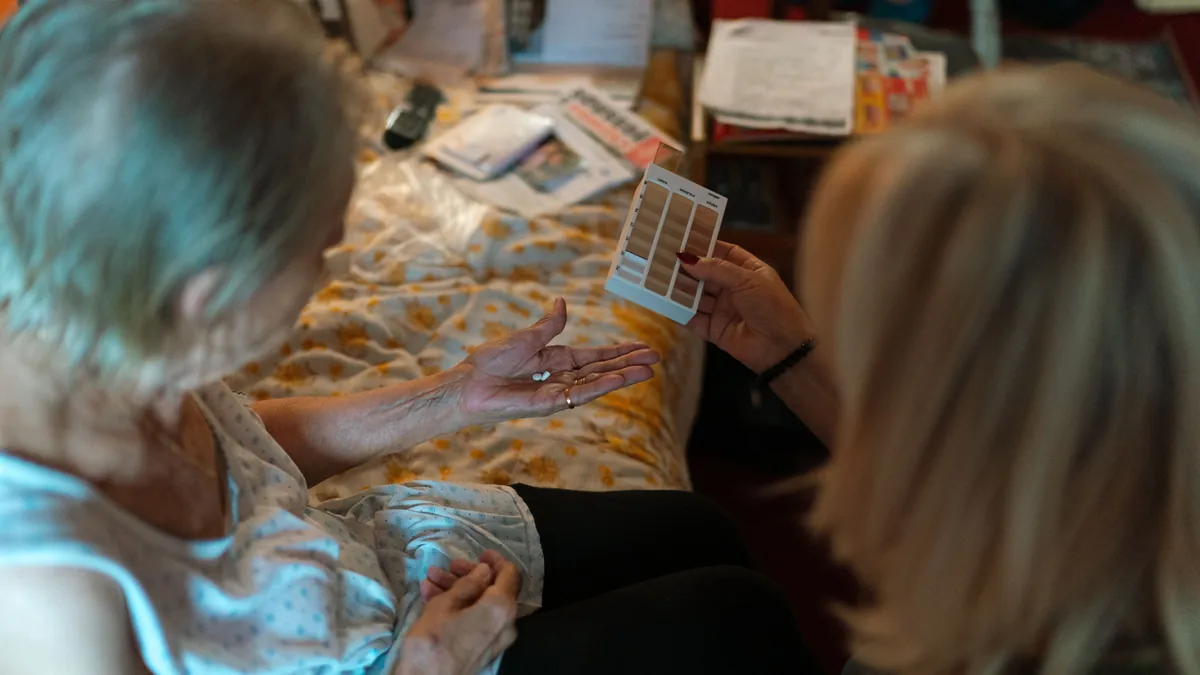 A caregiver helps an older woman sort her medications with a pill organizer at her bedside