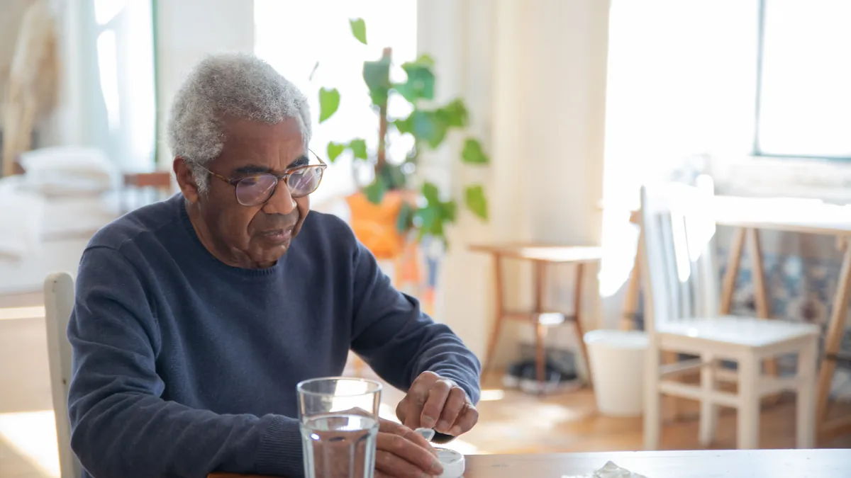 An older man at his kitchen table takes his daily medication with a glass of water