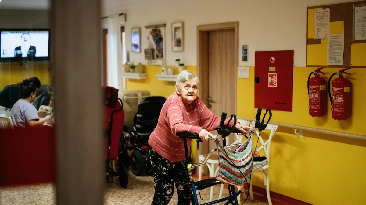 An older woman walks with a rolling walker through a care facility, where staff watch for wandering risk